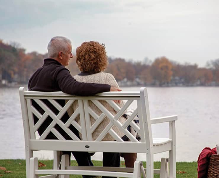 A couple with gray hair sits closely on white outdoor furniture, overlooking a calm lake surrounded by autumn trees.