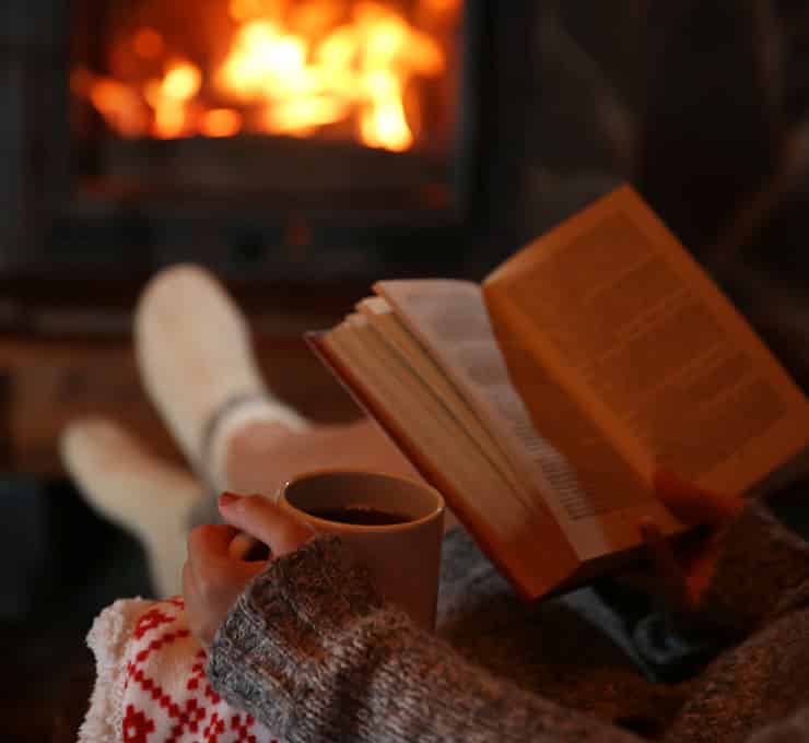 A person reads a book and holds a cup while seated near the hearth & fireplace in Cumberland, MD. Warm socks and a blanket are visible.