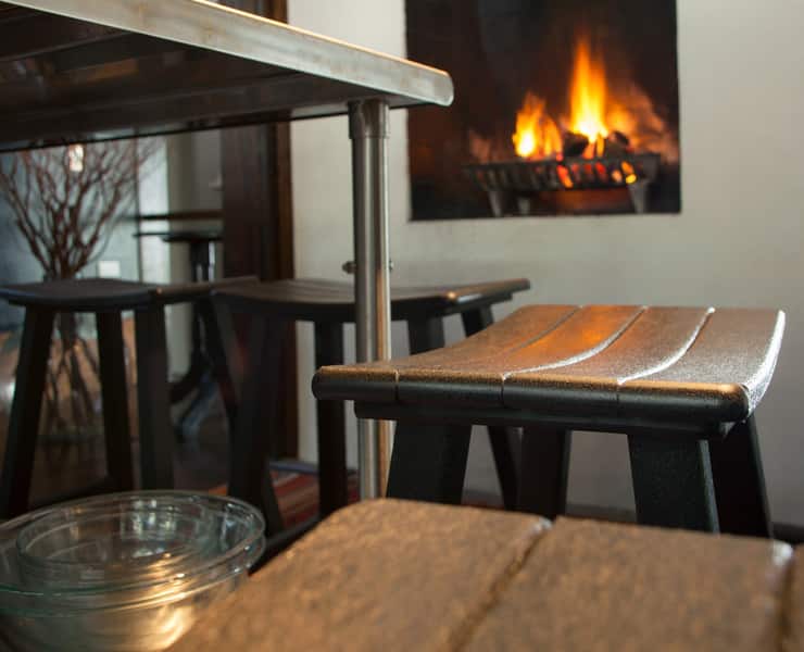 A close-up view of a cozy indoor space featuring a table and stools reminiscent of outdoor furniture in the foreground, with a lit fireplace in the background.