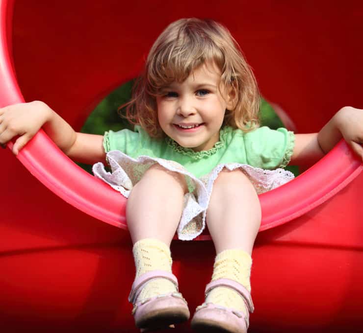 A young girl in a green dress and yellow socks smiles while sitting inside a red circular play structure, part of one of the many outdoor playsets, holding onto the edges with both hands.