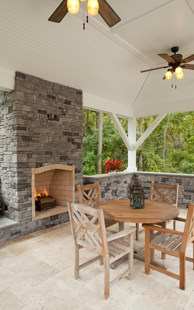 Covered outdoor patio with stone fireplace, round wooden table, four matching chairs, two ceiling fans with lights, and a view of green trees in the background.