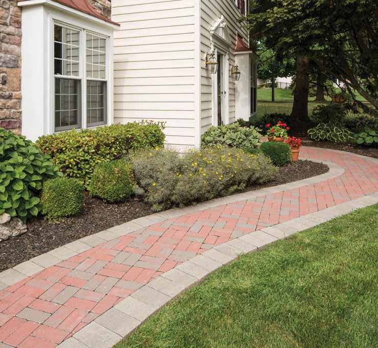 A brick pathway curves along the edge of a landscaped yard with green shrubs and plants, showcasing a beautiful mix of hardscape materials as it leads to the side entrance of a house with white siding and stone accents.