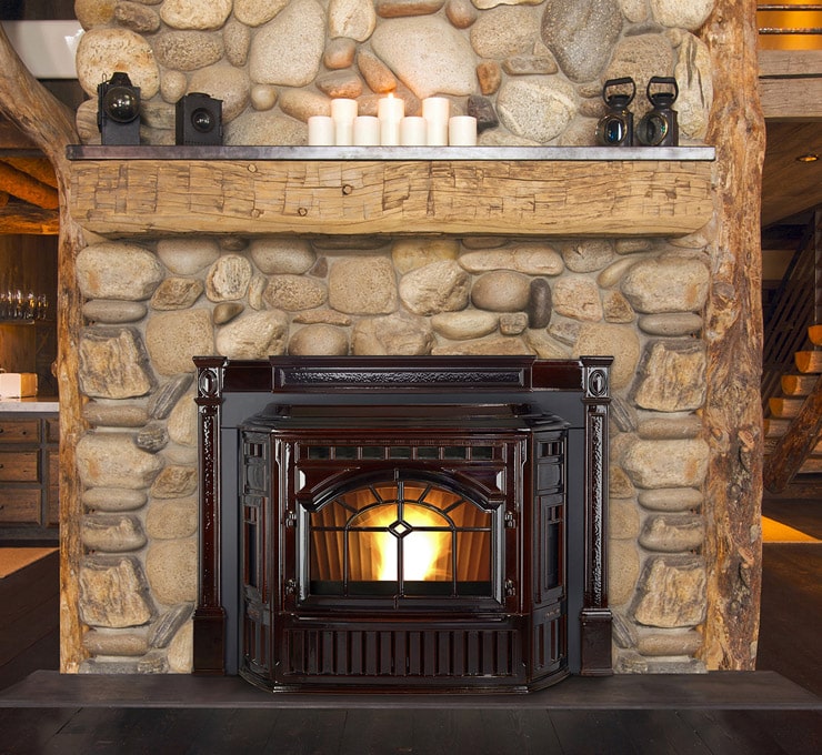 A hearth & fireplace Cumberland MD boasts a stone fireplace with a rustic wooden mantel displaying candles, housing a dark wooden stove. The stonework, comprised of variously sized fieldstones, enhances a cozy, rustic ambiance.