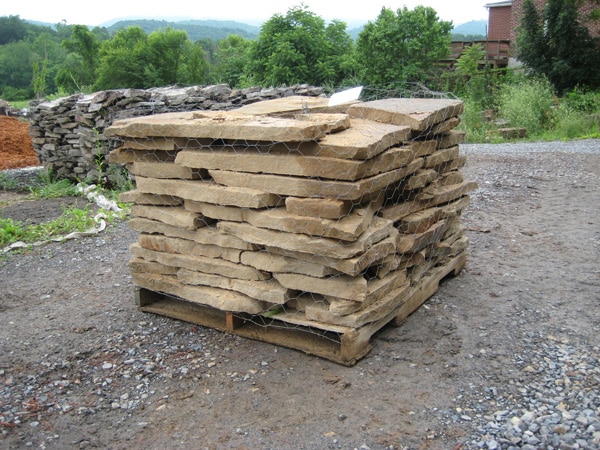 A stack of large, flat stones is piled on a wooden pallet outdoors, surrounded by greenery and other stacks of stones.