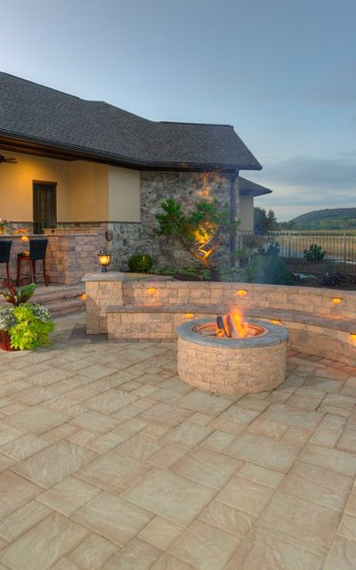 A stone patio with a circular fire pit, benches, and adjacent house, features warm lighting and a distant view of hills at dusk.