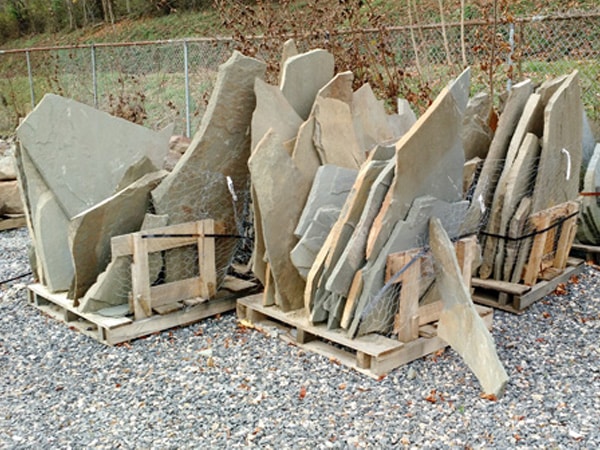 Two wooden pallets hold large, irregularly shaped stone slabs in various shades of gray, placed outdoors on a gravel surface near a chain-link fence.