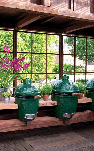 Three Big Green Egg ceramic grills are placed on a wooden counter inside a greenhouse with potted plants and flowers in front of large windows.