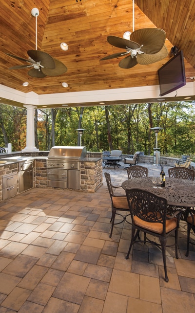 A covered outdoor kitchen with stainless steel appliances, wooden ceiling, two ceiling fans, and a stone patio. A dining table with chairs and a seating area are also present.