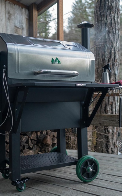 A silver and black outdoor grill on a wooden porch, with smoke coming out of the chimney. Firewood is stacked in the background.