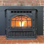 A lit fireplace insert with a black metal frame is set against a red brick wall. The flames are visible through the glass front, providing a warm, glowing ambiance.