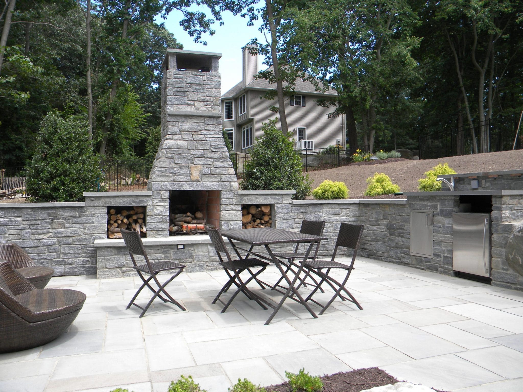 Outdoor patio with stone fireplace, dining table with four chairs, adjacent lounge chairs, built-in grill, and surrounding greenery and trees. A house is visible in the background.