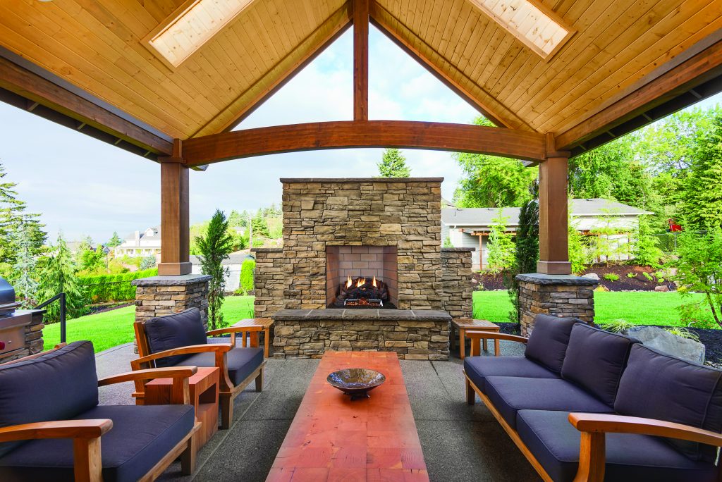 A covered outdoor patio with a wooden ceiling and stone fireplace, featuring wooden furniture with blue cushions and a wooden coffee table, set in a green backyard.