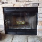 A stone fireplace with a black metal frame and glass doors. Inside, there's a yellow log starter positioned on the grate. Firewood and a fireplace tool set are visible beside the fireplace.