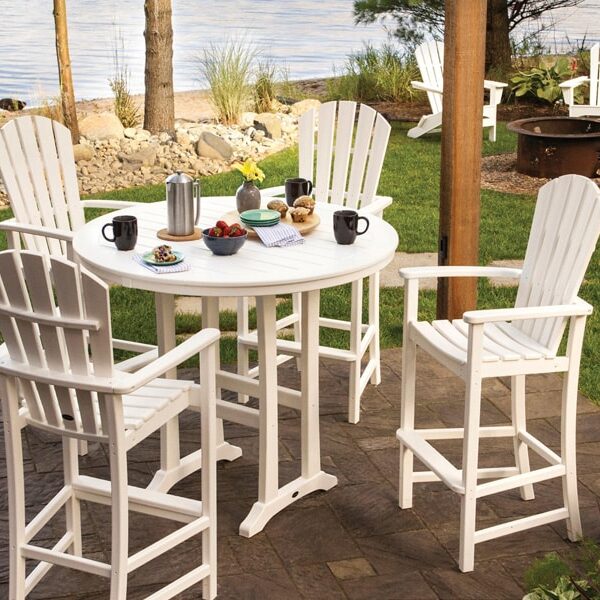 A white outdoor dining set with four chairs, a round table holding coffee, pastries, and fruit, is set on a patio near a grassy area facing a lake. In the background are more chairs by a fire pit.