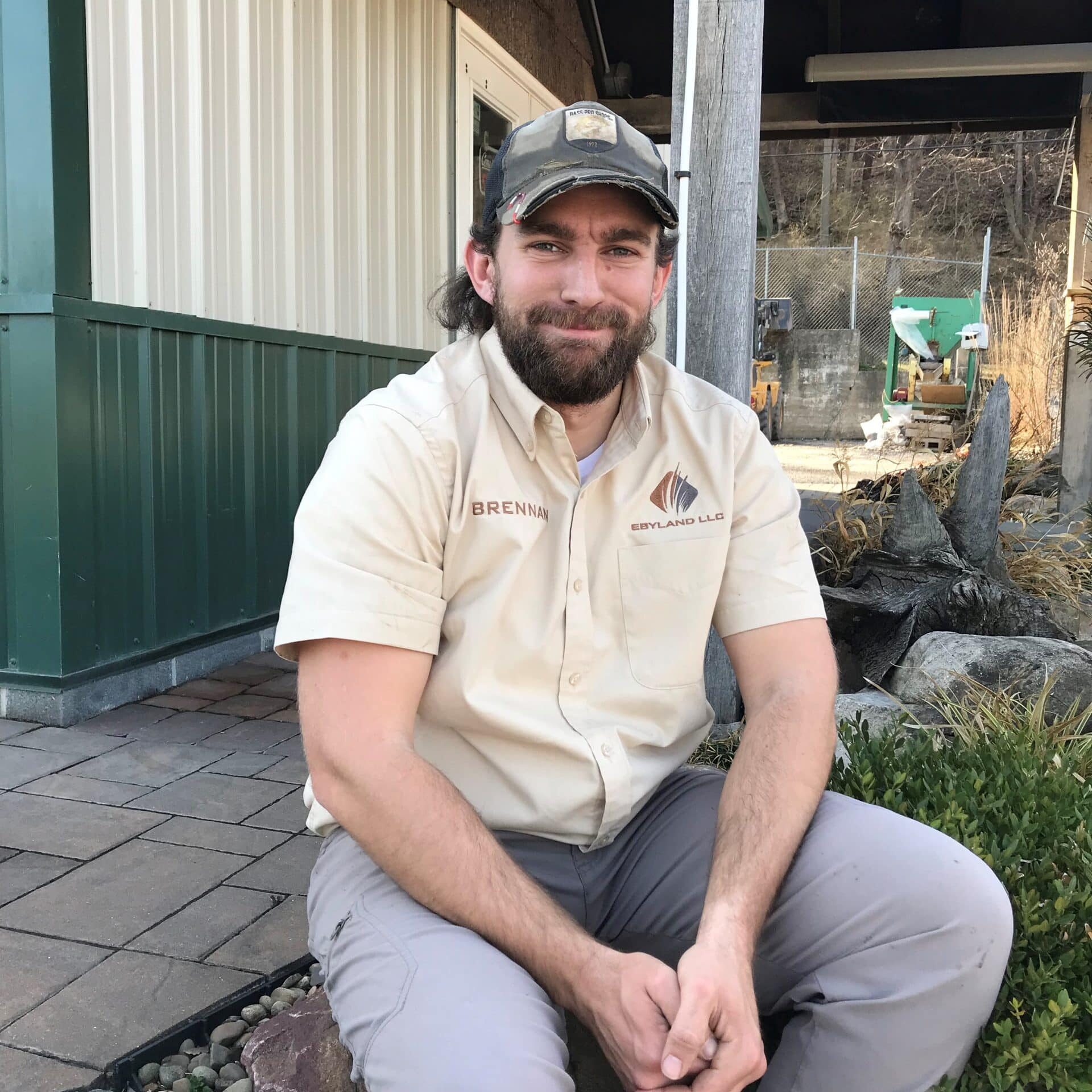 A man wearing a beige shirt and cap with "Brennan" on his shirt is sitting on the edge of a landscaped area with rocks, in front of a building with green and white siding.