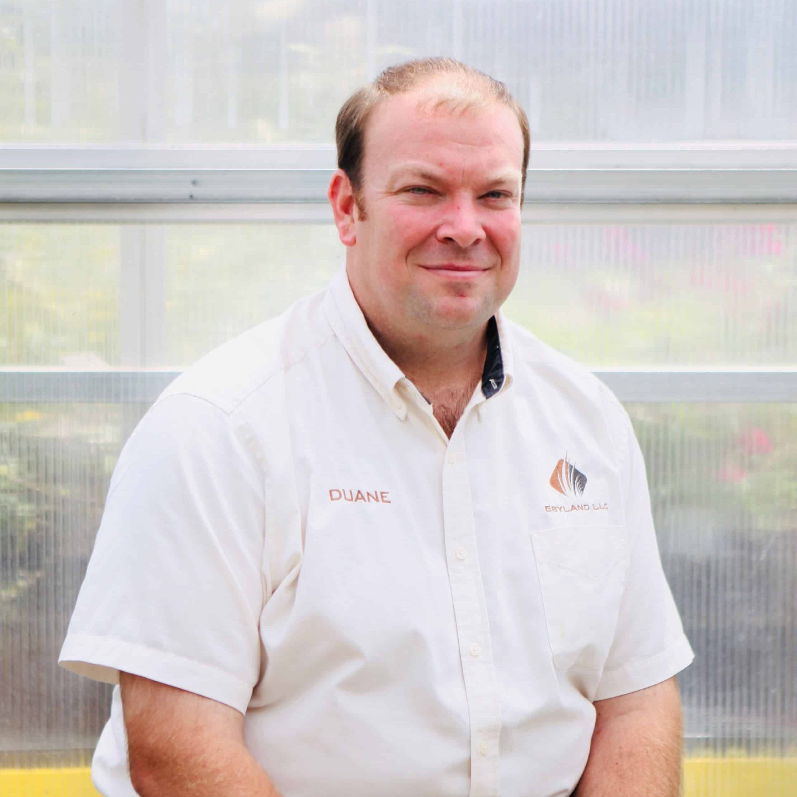 A man with short hair is seated, wearing a white collared shirt with the name "Duane" and a company logo embroidered on it. He is positioned in front of a translucent background.