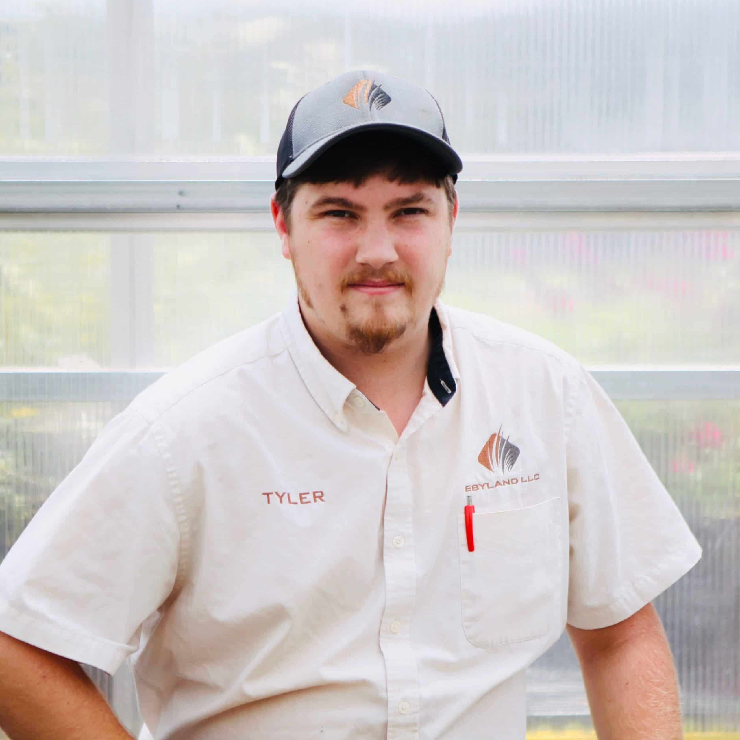 A man in a white shirt and baseball cap with an orange and black logo stands indoors. The shirt has "Tyler" embroidered on it.