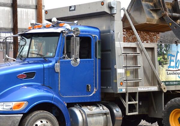 A blue dump truck is being loaded with gravel by a front loader. The truck bears a company logo for landscaping services.