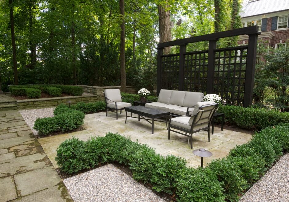 An outdoor seating area with a black metal lattice screen, cushioned chairs, a sofa, and a coffee table on a stone patio surrounded by greenery and trimmed hedges is perfect for designing a beautiful backyard.